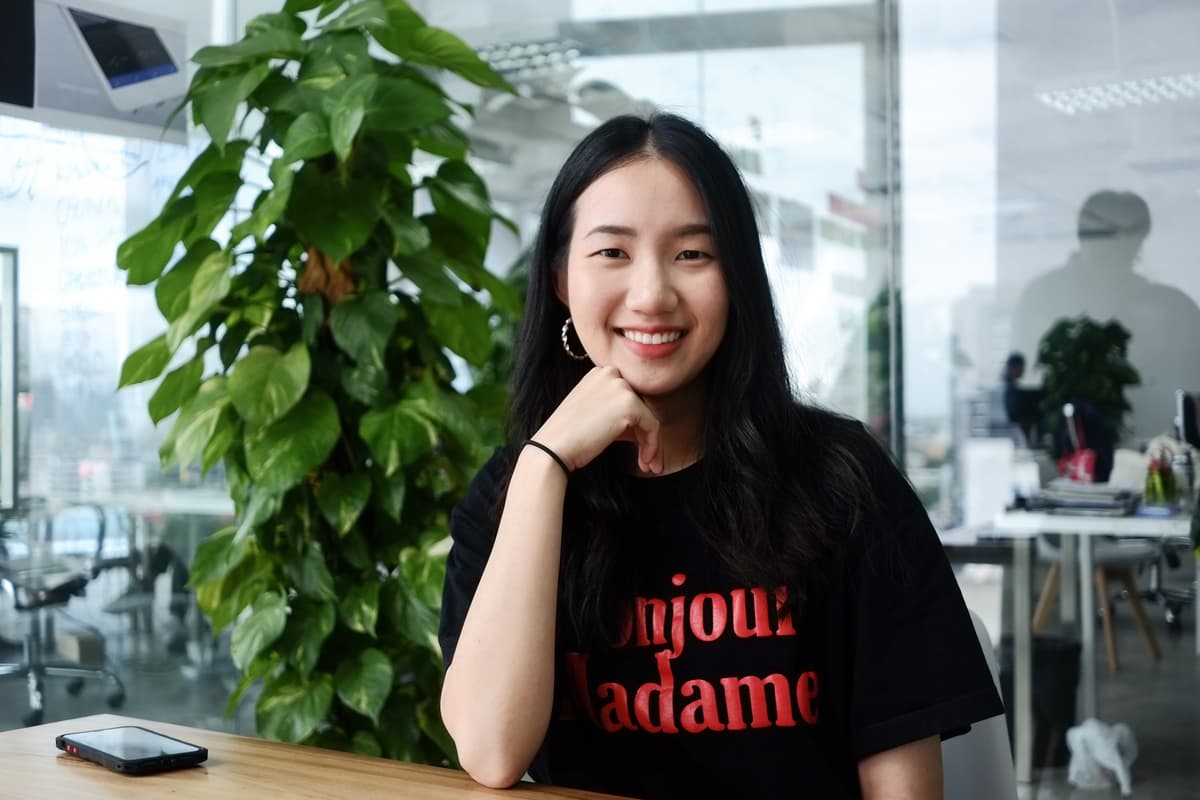 พนักงานนั่งยิ้มที่เคาน์เตอร์ไม้ในออฟฟิศเปิดโล่ง มีต้นไม้ห้อยและผนังกระจกกั้นห้องประชุมด้านหลัง